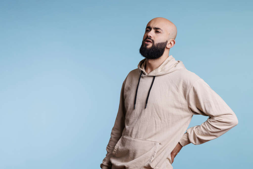 A bald but bearded man in his twenties or thirties, is holding his lower back and his face expresses discomfort. The picture is in an article relating to online physiotherapy and how it can help treat musculoskeletal conditions