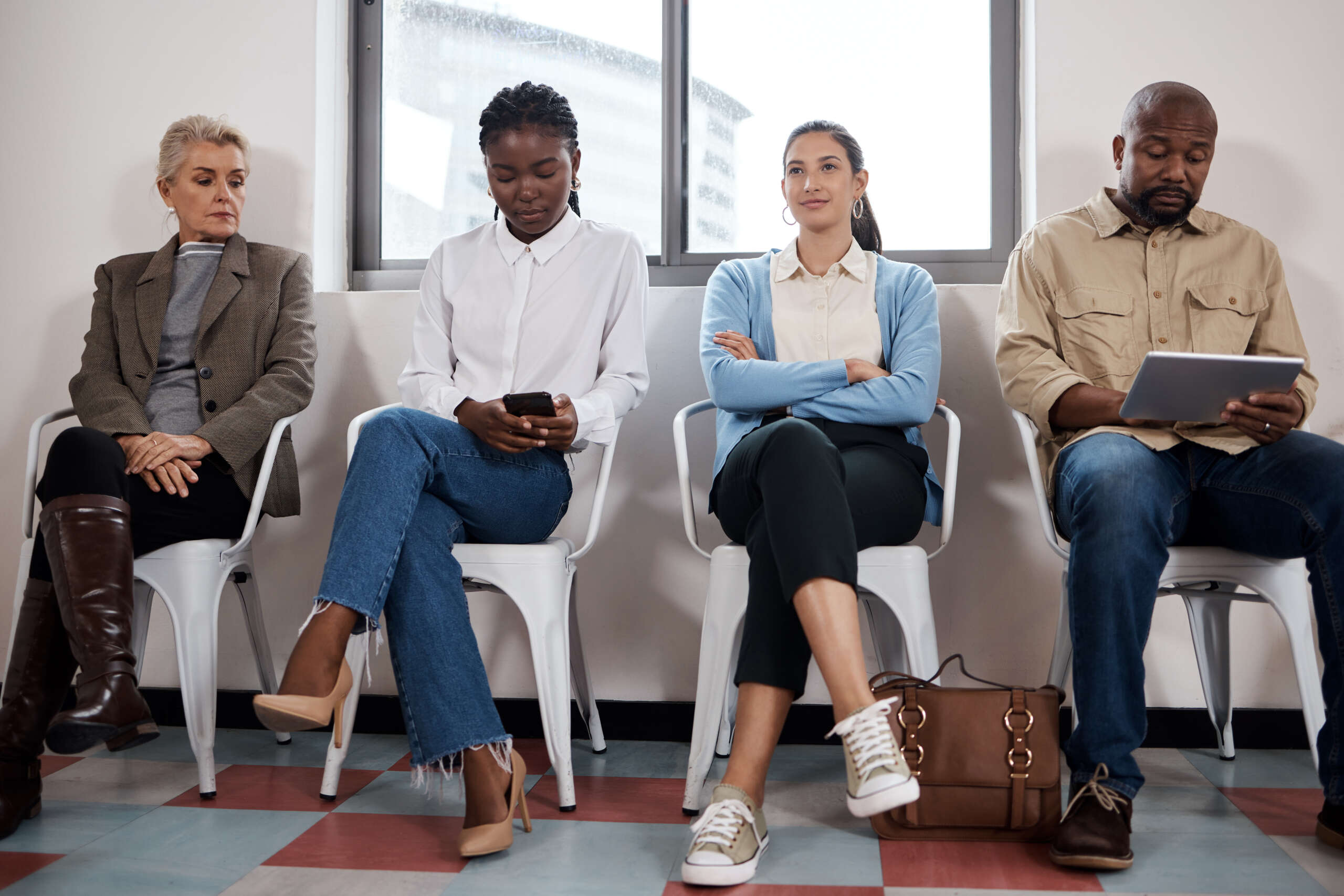 A waiting room with 4 people, they are waiting for a physiotherapy appointment. We can only see a small part of the room implying that it is very busy. One patient is on a tablet and one on a phone. Only one is smiling.