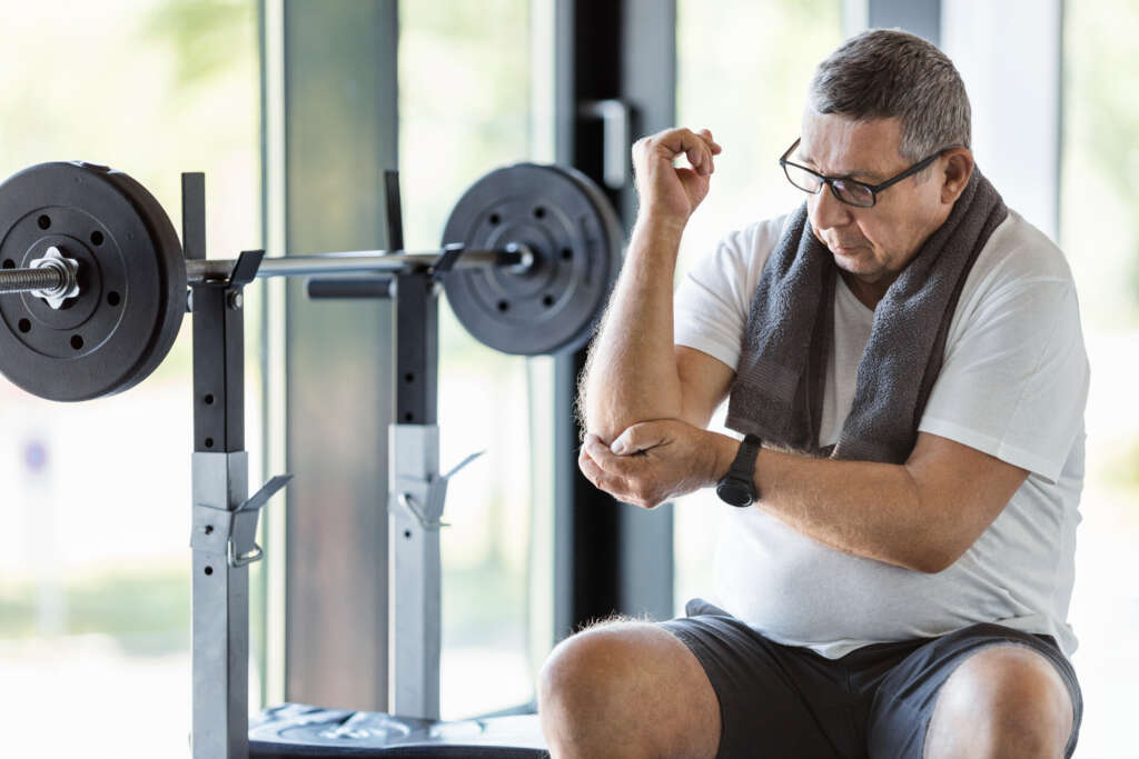An active senior man is sitting in a gym holding his arm as if suffering from tennis elbow pain, the image is in an article about tennis elbow and how physiotherapy can help this condition. The man is sitting near some weights and has a grey towel around his shoulders. He is wearing a white t-shirt.