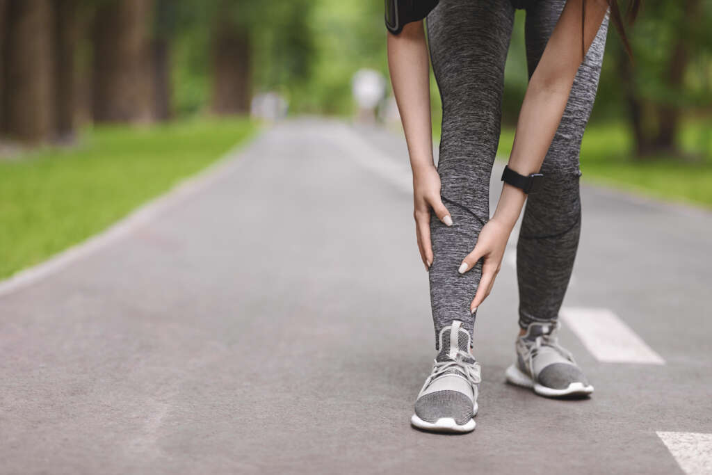 Lower limbs of a runner who appears to be young female / non binary runner suffering from shin splints while jogging in park, massaging painful area. Image is a header to a blog post about the condition shin splints.