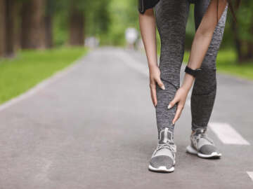Lower limbs of a runner who appears to be young female / non binary runner suffering from shin splints while jogging in park, massaging painful area. Image is a header to a blog post about the condition shin splints.